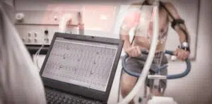 Foreground, researcher watches heart rate readings on a monitor. Background, person taking a stress test. 