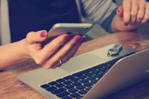 Close-up picture of a woman's hands holding an iPhone and hovering near a computer keyboard