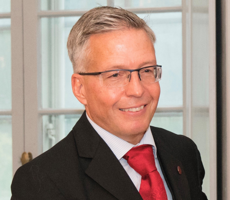 le Dr Bergquist porte des lunettes, un costume et une cravate rouge.