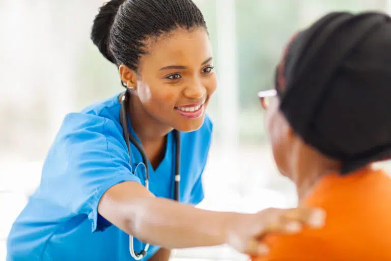 The image depicts a healthcare setting where a young female healthcare worker, wearing a blue scrub top and a stethoscope around her neck, is holding the hands of an older patient.