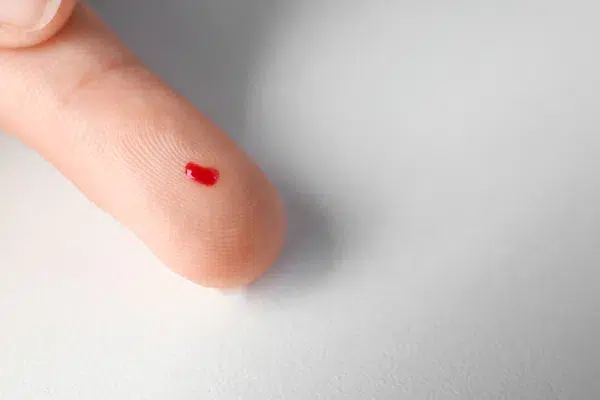 a close-up of a finger with a drop of blood on it, against a white background.