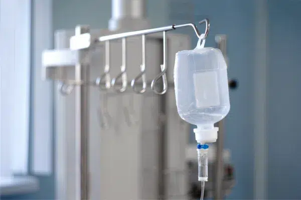 L'image montre une poche de perfusion, attachée à un support métallique, indiquant un environnement médical ou clinique.
