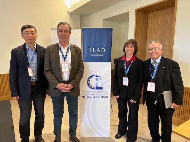 Photo de Linda Tannenbaum, Jonas Bergquist, David Systrom et Wenzhong Xiao à côté d'une bannière pour la première conférence internationale sur l'EM/SFC et la COVID longue au Portugal.