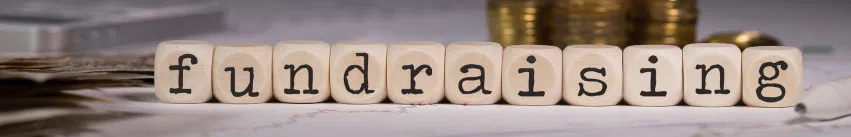 The image shows the word "fundraising" spelled out using wooden blocks with black letters. In the background, there are stacks of coins and some documents.