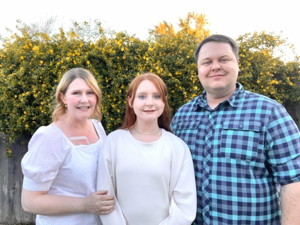 Cette image montre Wendy et sa famille debout ensemble à l'extérieur devant un buisson fleuri. Wendy porte un haut blanc, sa fille porte une chemise de couleur claire, et son mari porte une chemise à carreaux bleue.