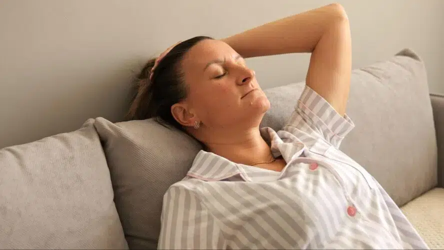 The image shows a woman with ME/CFS resting on a couch. She is wearing striped pajamas and leaning back with her eyes closed.
