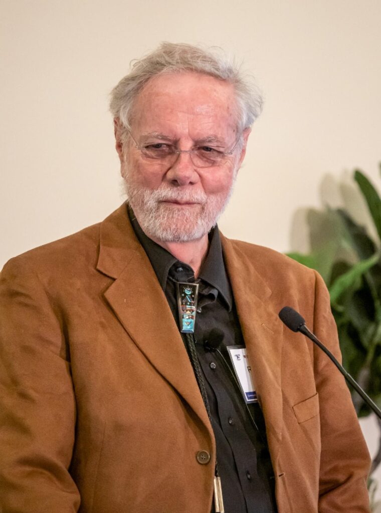 Photo du Dr Ron Davis debout devant le micro. Il porte un blazer marron chaud sur une chemise noire.